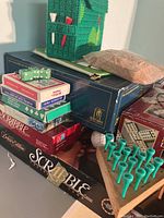 Stacked boxed board games and game components including Scrabble Deluxe Edition box, Trivial Pursuit box, dominoes box, dice, and green plastic golf tees.