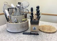 Photo of J.A. Henckels International knife block with knives and cutting tools, gray spinning utensil holder with various kitchen utensils, and two woven hot pads on a countertop.