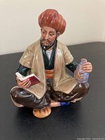 Front angle showing the seated figure holding a red book and striped jug, wearing a brown outfit and turbans.