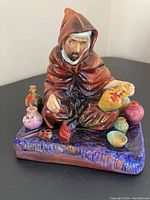 Front view of The Potter porcelain figurine showing the figure in a reddish-brown cloak with pots and bowls on a purple base.