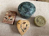 Four small containers on carpet surface; two rectangular marble-like, one heart shaped, one round carved stone with elephant, and one pottery container.