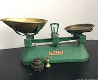 Full view of the green Timothy White & Taylor Ltd kitchen scale with two brass weighing pans and weights placed in front.