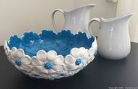 Photo showing the blue interior and white and blue daisy exterior design of the ceramic bowl with scalloped edges, with two white ceramic pitchers in the background.