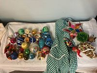 Overview photo showing about 31 colorful vintage Christmas ornaments, a green and white retro apron, a multicolored glass star decoration, two mistletoe balls with elves, and a small green plant.