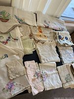 Various sizes and styles of hand embroidered vintage linens including scarves, tablecloths, napkins, and doilies arranged on a black surface.