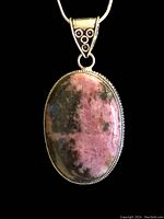 Front view of oval rhodonite pendant set in silver filigree with attached silver chain on black background.