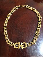 Full view of chunky gold plated chain choker necklace laid flat on a wooden surface. Shows interlocking horseshoe style clasp detail in gold tone metal.
