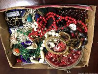 Top down view of a box filled with mixed vintage costume jewelry including beaded necklaces, bracelets, earrings, and a red sequin pouch.