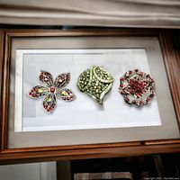 Three costume jewelry brooches with red and green rhinestones displayed in a frame.