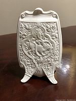 Front view of the small French bisque vase showing detailed embossed floral scrolling and heart-shaped central motif on two feet.