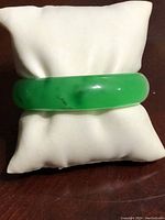 Solid green jade bangle bracelet displayed on white cushion, showing smooth surface and natural darker inclusions.