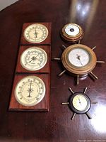 All five vintage weather instruments arranged on a wood table showing variety of sizes and styles, three mounted to a wooden panel and two ship wheel styled pieces.