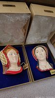 Two hand-painted Bossons easel-mounted head plaques of Catherine of Aragon and Anne Boleyn displayed upright on blue velvet with gold trim, with original packaging in the background.