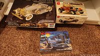 Three boxed model car kits including Monogram Packard Boattail Speedster, AMT Ford '29 Model A, and Elvira Macabre Model car on carpet.