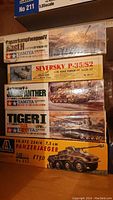 Five unassembled plastic model kits stacked, showing Tamiya and Italeri military vehicle kits and Williams Bros aircraft kit.