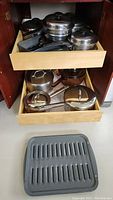 Wide view of two drawers in a kitchen cabinet filled with various stainless steel pots and pans with lids and handles. Below drawers is a cast iron style broiler/grill pan.