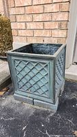 Single greenish deep square resin planter with lattice design on all sides, placed on ground by brick wall. Visible wear and damage.