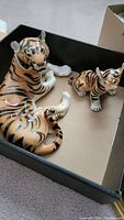 Two porcelain tiger figurines in a box: one adult lying down, one cub standing.