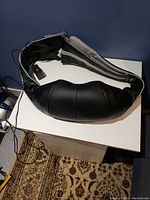 Top view of black ergonomic back massager placed on white table, showing padded design with mesh fabric central panel and cable attached.