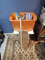 Orange bin filled with vinyl records, DVDs, basket, and a wood tool; cardboard sheets in front with a measuring ruler indicating about 16 inches height