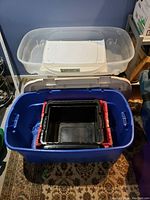 Photo shows multiple plastic storage bins stacked including a large blue bin holding smaller black and red crates, a large transparent storage bin at back.