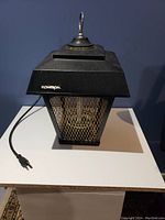 Front view of black Flowtron bug zapper with attached power cord on white table and blue background.