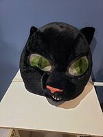 Front view of the large black plush cat head costume showing green eyes, a pink nose, and teeth.