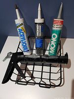 Photo of three tubes of caulking and one black caulking gun on a metal rack with blue backdrop