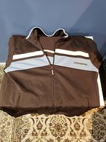 Photo of the folded brown Converse warm up sweater with white and light blue stripes, zippered front, showing logo detail on chest.