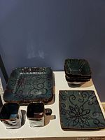 Overall view of the ceramic dishware set showing square plates, bowls, and cups with blue-green glaze and dark floral patterns.