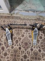 Double bass pedal laid flat on patterned carpet, showing both footboards connected by metal linkage rods with Mapex logos visible on footboards.