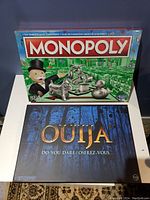 Photo showing two unopened board games: Monopoly and Ouija, stacked on a table against a dark wall.