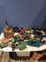 Overview of collection of military-themed GI Joe toy vehicles spread on a white table including several tanks, helicopters, a boat, and a large yellow tracked vehicle with a dome.