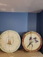 Two round decorative hat boxes side by side with rope handles, one with a teddy bear and honey design and the other with purple irises and floral border.