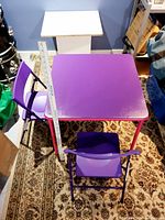 View of kids purple table with two purple plastic children chairs placed on a patterned floor.