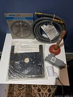 Lot items displayed on white surface including circular saw blade in package, router base plate sealed in plastic, coiled grey plumbing hose, red plunger, and small gray tool part.