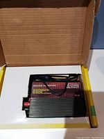 Open box showing the black power inverter unit with cooling fins and red and black terminal connectors.