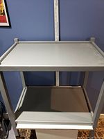 Front view of the grey two-shelf rolling cart against a blue wall.