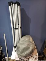 Collapsed white metal tent frame standing upright against a blue wall.