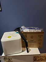 White fabric steamer unit with control knob and attached hose with nozzle seen from the front with accessories box behind