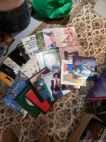 Photo showing a collection of colorful vinyl record covers laying on a patterned carpet, featuring various artists including Olivia Newton-John and Pat Benatar.