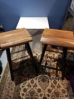 Two wooden stools with polished light natural wood seats and black tubular metal legs placed on patterned carpet with blue wall background.