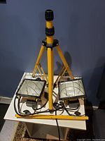 Overall view of the yellow tripod holding two rectangular work lights with metal protective cages, showing cords and setup on a white surface.