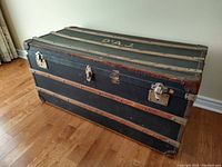 Front side view of the large vintage trunk showing black exterior, wood slats, leather corner reinforcements, and metal locking hardware.