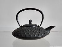 Black cast iron tea pot or sake warmer with hobnail pattern, curved spout, round lid with knob, and a high curved handle viewed from the side.