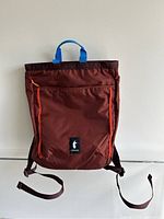 Front view of the maroon Cotopaxi backpack showing blue carrying handles, orange piping, front zippered pocket, shoulder straps, and logo patch.