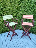 Two tall folding director style patio chairs, one with light green fabric and one with faded coral pink fabric, wooden frames and armrests. Chairs placed on a blue deck in front of greenery.