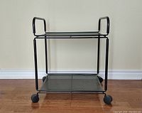 Front view of the black metal serving cart with two perforated shelves and four wheels.