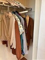 View of the vintage women's clothing hanging on a rod showing multiple tops and jackets in various colors from cream, brown, floral prints, teal, to light blue.