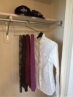 White men's button-up shirt hung on a metal rack with four silk ties of various colors and patterns hanging beside it. Two hats rest on top shelf above them.